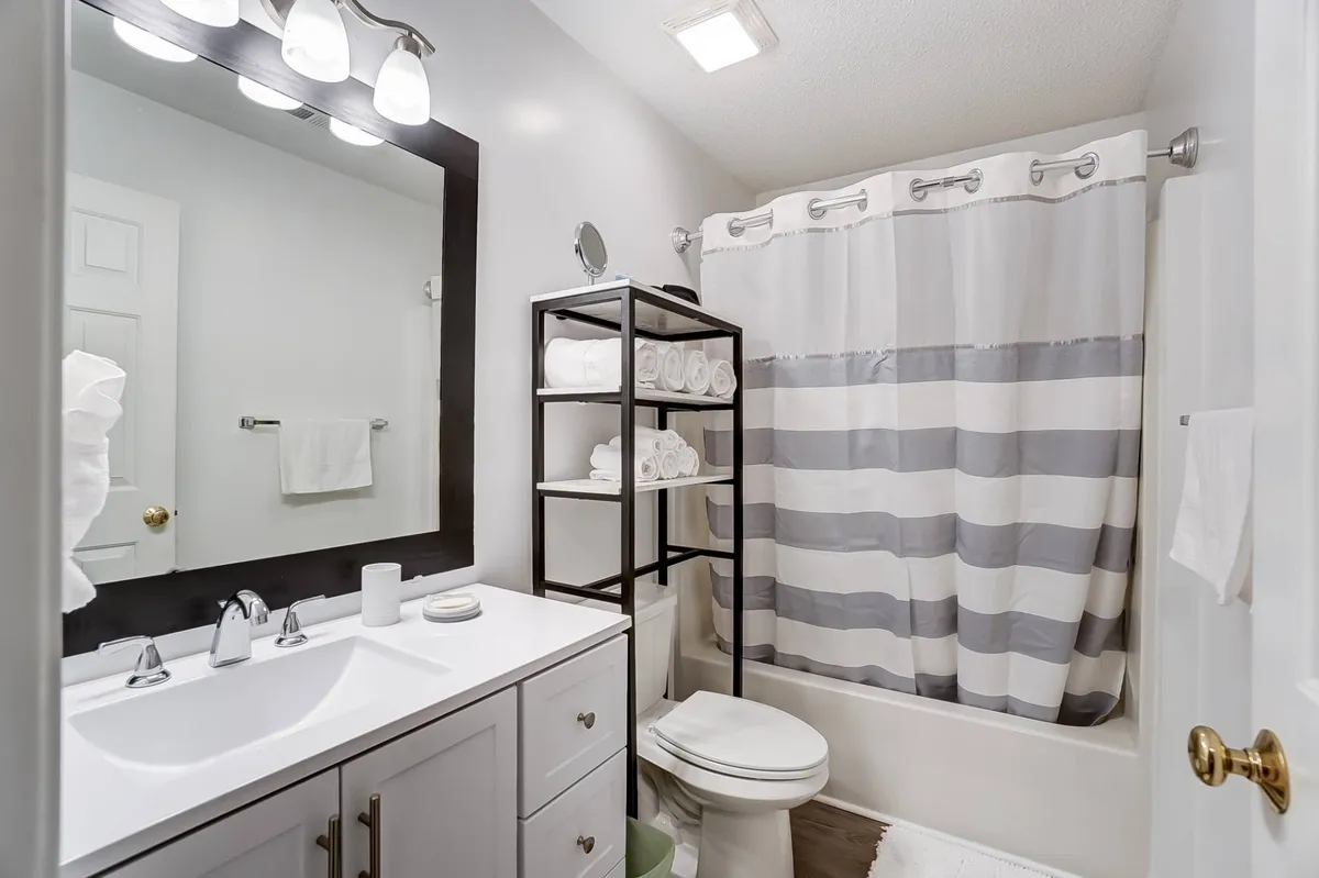 A bright, fresh bathroom featuring a vanity with dual sinks, a wide framed mirror, a striped shower curtain, neatly organized towels, and a storage rack. The overall vibe is clean and inviting, perfect for unwinding or freshening up after a busy day.