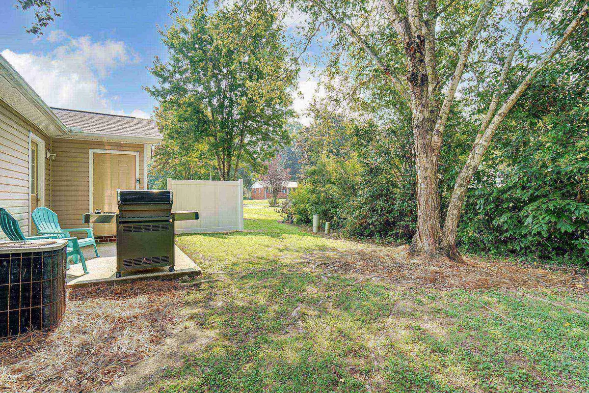 A pleasant backyard patio area with a grill, outdoor seating, and lush greenery. The scene evokes a casual, friendly mood, with plenty of space for family or pets to enjoy the outdoors, making it ideal for barbecues or afternoon relaxation.