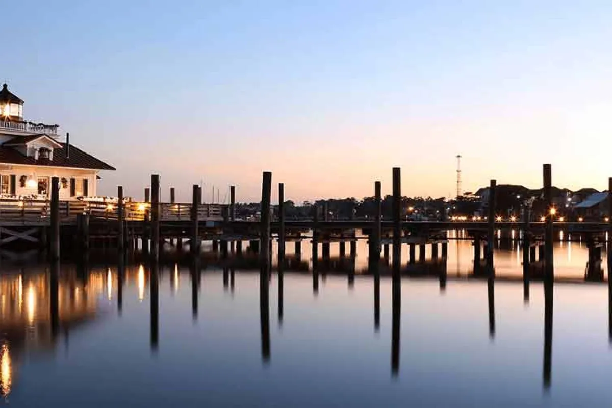 A serene waterfront scene featuring a dock, calm water with mirror-like reflections, a nearby building, and a colorful sky. The setting is tranquil and picturesque, suggesting opportunities for relaxation and leisure after work.