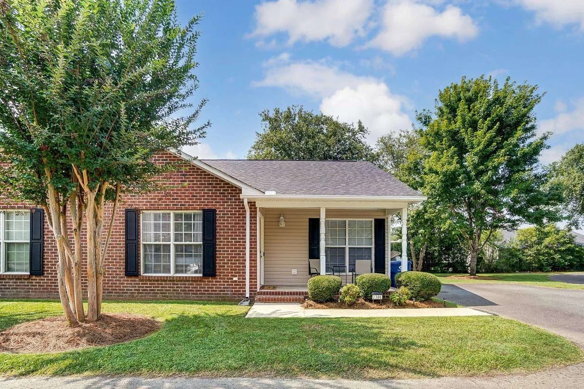 The hero image showcases the charming brick facade of the Bulldog Bungalow, framed by classic window shutters, a welcoming front porch with seating, and a beautifully landscaped yard. The porch invites relaxation, while the lush greenery creates a sense of privacy and tranquility, making the entryway warm and inviting.