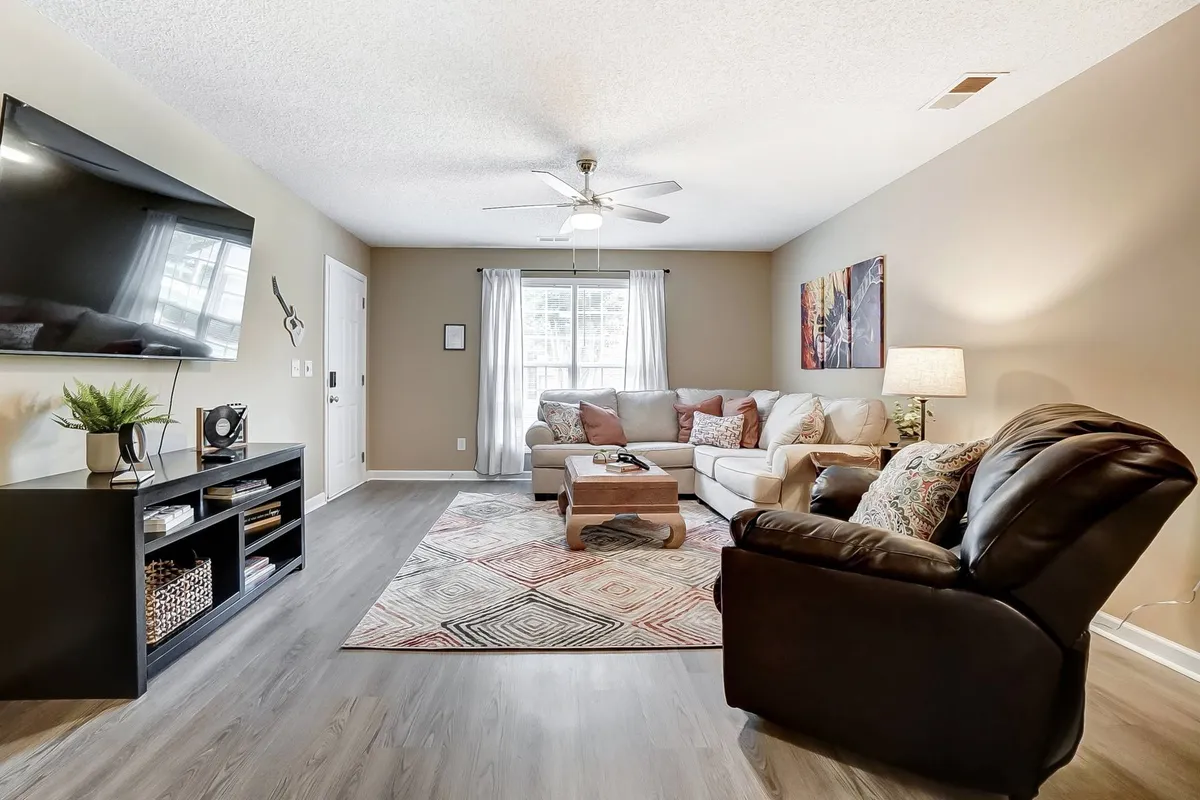 A comfortable living room featuring a plush sofa, coffee table, area rug, and a large television. Natural light streams in through a nearby window, creating a warm and inviting atmosphere ideal for relaxation, socializing, or group activities like watching TV together or reading.