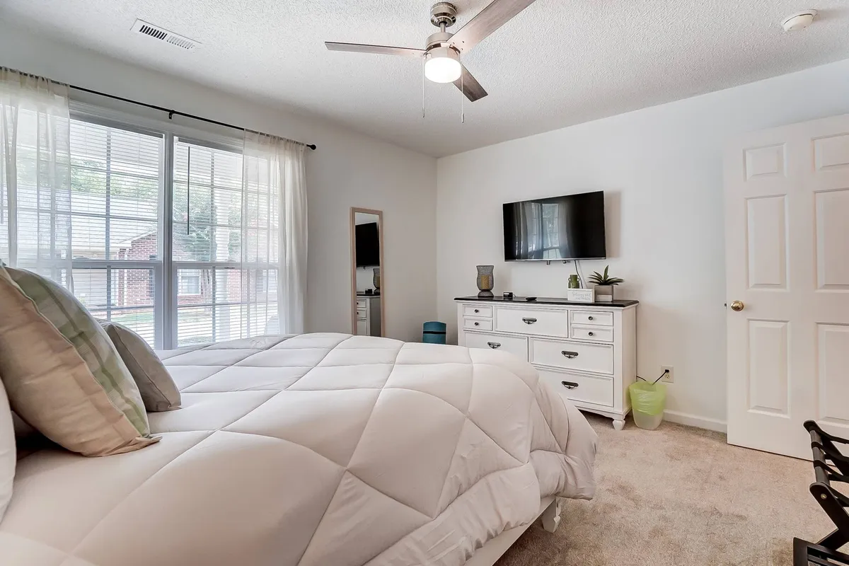 A serene master bedroom with a plush bed covered in a white comforter, a dresser topped with decorative items, a wall-mounted television for private entertainment, and a large window flooding the space with natural light. The atmosphere invites relaxation and comfort, ideal for restful nights.