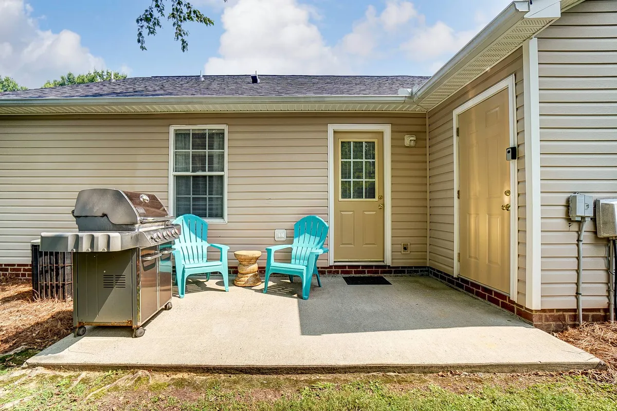 A private backyard patio featuring a barbecue grill, two blue chairs, and a small table. The setup is casual and perfect for outdoor dining or relaxing, with enough space for informal gatherings or solo moments. The fence in the background adds privacy, and the setting captures the inviting atmosphere of an outdoor retreat.