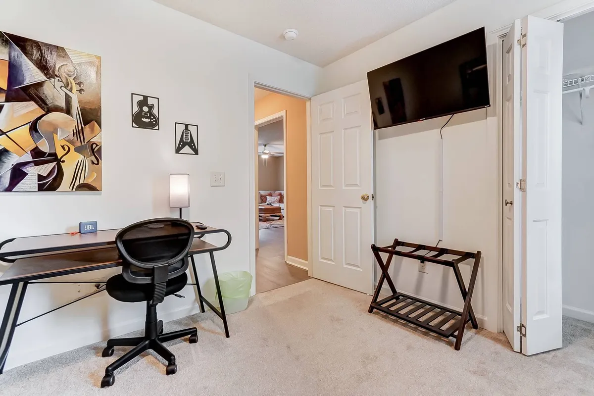 A thoughtfully arranged workspace featuring a desk with a comfortable chair, stylish abstract artwork, and a wall-mounted TV. The setup suggests a versatile environment, suitable for both productivity and relaxation, with ample lighting and a sense of modern comfort.