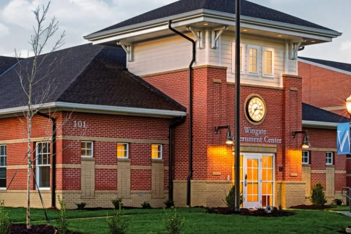 A welcoming brick building with a prominent clock tower and well-maintained landscaping, representing a central community hub or government services space. The entrance doors and inviting pathway create an approachable connection to the local area.