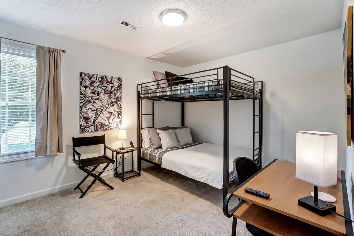 A warm and welcoming second bedroom featuring a full bunk bed, a practical desk for study or reading, wall art adding character, and a bedside lamp. The space is cozy and inviting, suitable for families or friends sharing the room.